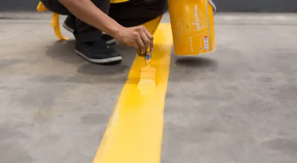 Persona pinta una línea amarilla en suelo de cemento sin pulir, usando una brocha y un cubo de pintura.