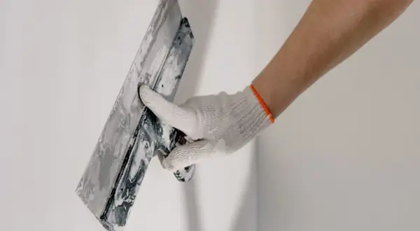 Mano enguantada aplicando material para revocar pared con llana.
