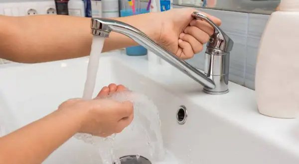 Mano ajustando el grifo para aumentar la presión del agua mientras se lava las manos en un lavabo blanco.