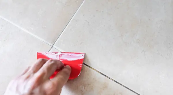 Mano aplica masilla con espátula roja para reparar una junta de baldosas.