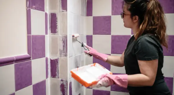 Mujer pinta pared con rodillo, cubriendo la cerámica esmaltada morada y blanca con pintura blanca.