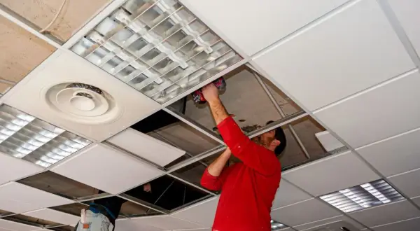 Un hombre con camisa roja instala un panel en **techos registrables**, junto a luminarias y rejillas de ventilación.
