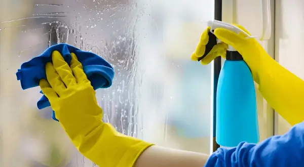 Manos con guantes amarillos limpian cristales muy sucios con un paño azul y spray.