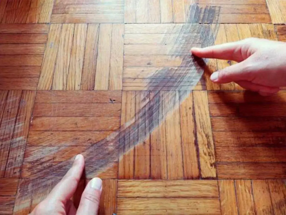 Manos señalan una mancha oscura en un suelo de parquet de madera. Se muestra cómo disimular manchas en parquet.
