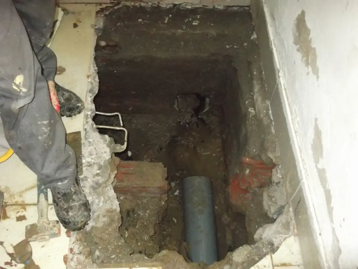 Excavación en el suelo de una vivienda para acceder a una arqueta con tuberías. Se ven restos de ladrillo y tierra.