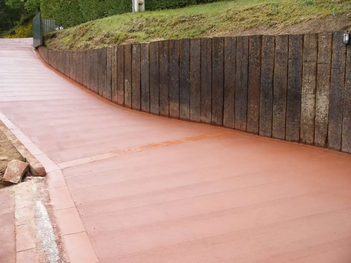 Camino de hormigón cepillado con borde de ladrillo, flanqueado por un muro de traviesas de madera.