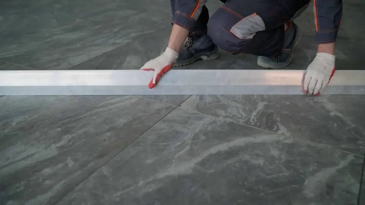 Man con guantes blancos y rojos coloca una regla metálica sobre azulejos rectificados, evitando problemas de desnivel.
