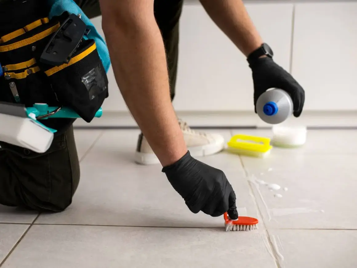 Persona con guantes negros usa un cepillo para limpiar juntas de azulejos, aplicando producto de limpieza.