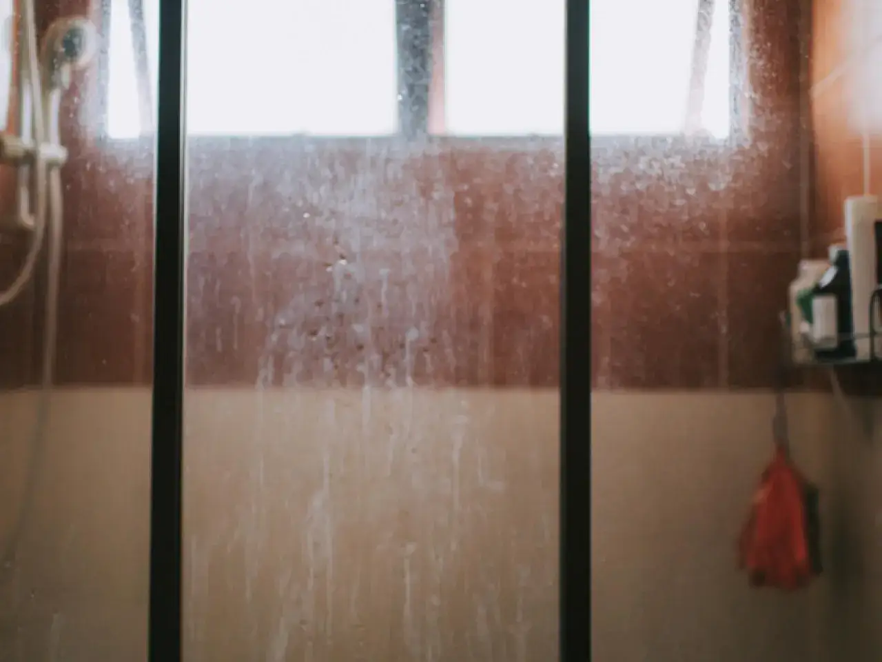 Mampara de ducha muy sucia con marcas de agua y jab&oacute;n. Se ven azulejos marrones y una escobilla roja colgando.