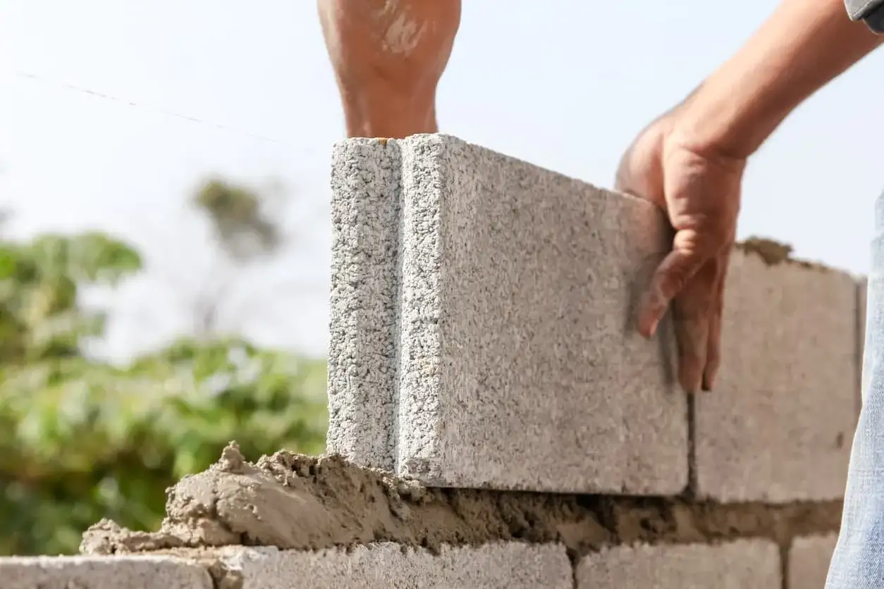 Manos construyendo un muro de bloques de hormig&oacute;n, colocando una pieza sobre cemento fresco.
