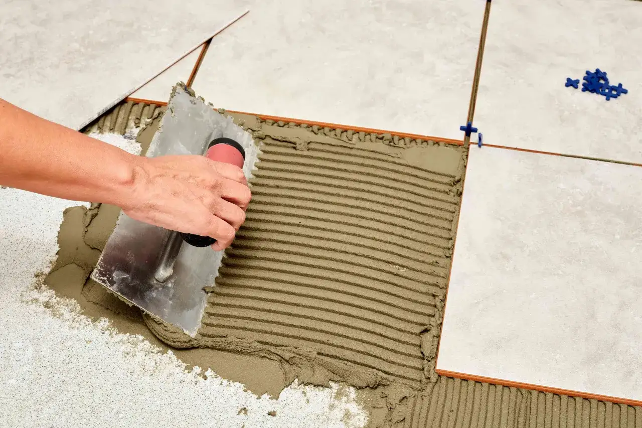Mano extendiendo cemento con llana para colocar baldosas. Se ven espaciadores azules y baldosas claras.