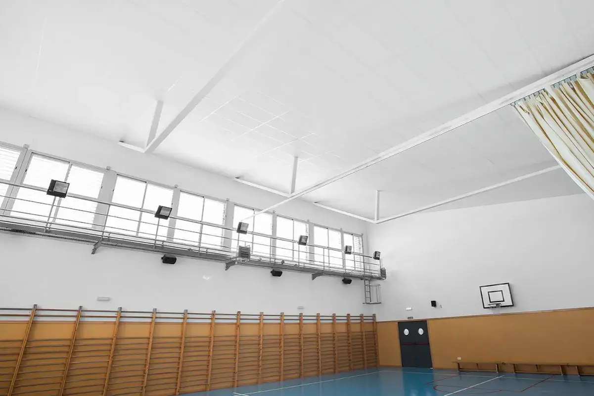 Techo de gimnasio con falso techo Heraklith, ventanas altas, canasta de baloncesto y pared de madera.