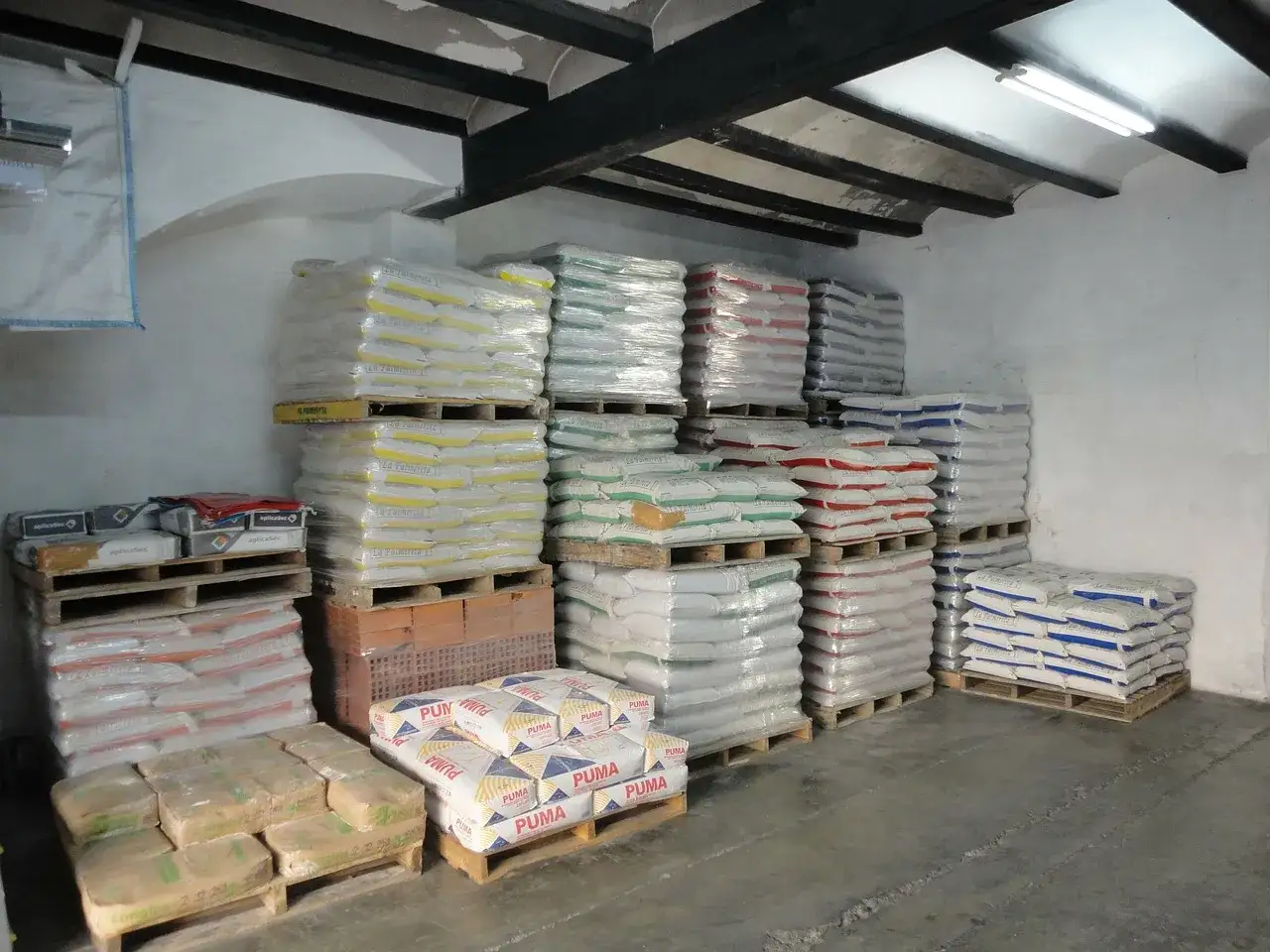 Almac&eacute;n con sacos de cemento y cemento portland apilados en palets. Se aprecian diferentes colores de sacos, indicando variedad de productos.