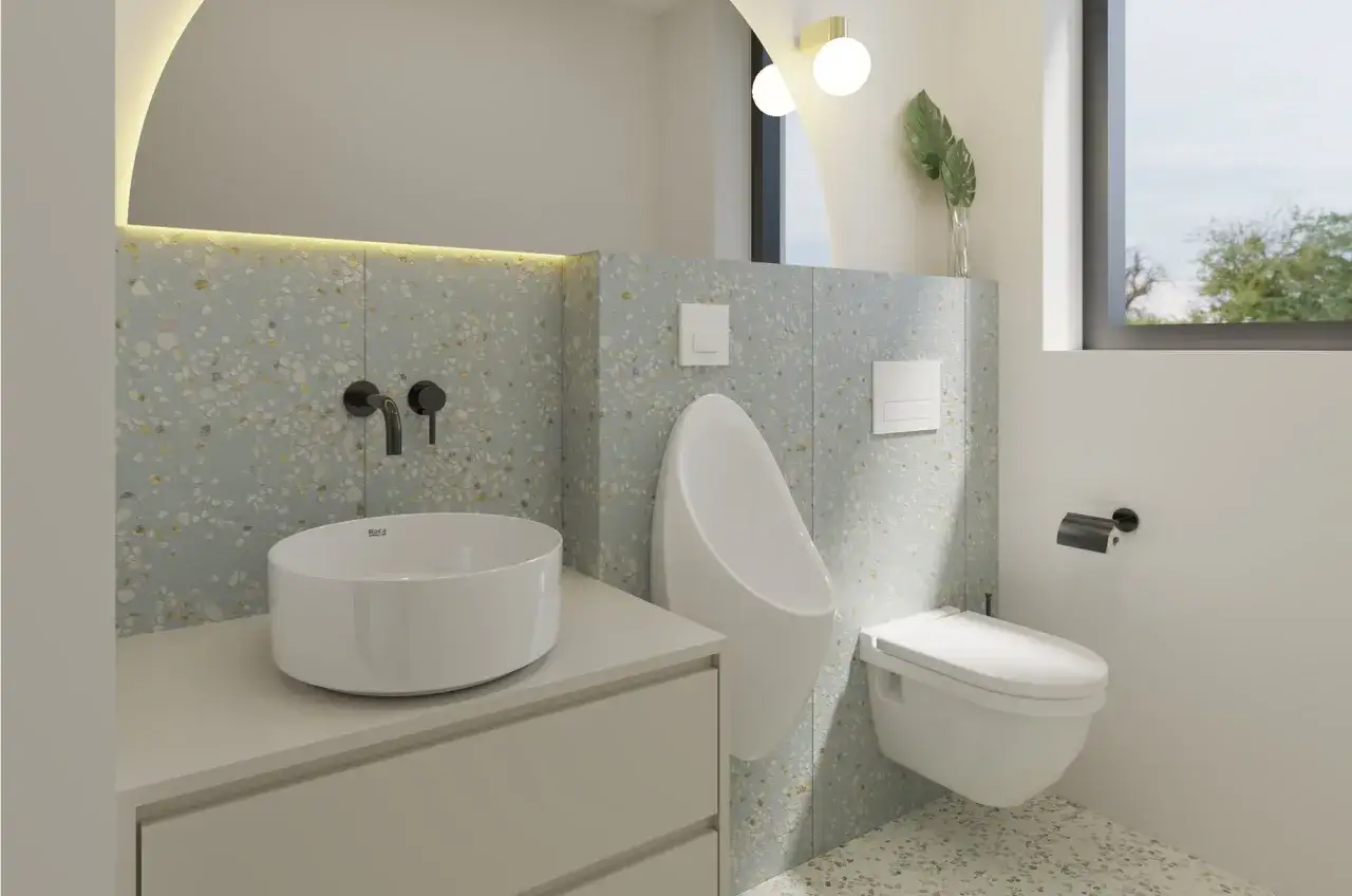 Ba&ntilde;os modernos con lavabo sobre encimera, urinario y WC suspendido. Las paredes presentan un dise&ntilde;o de ba&ntilde;os alicatados a media altura con un patr&oacute;n de terrazo en tonos pastel.