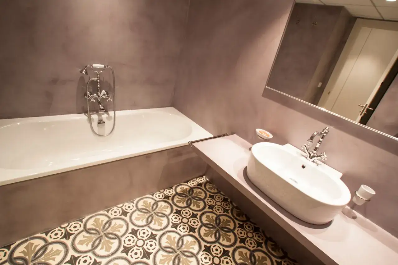 Elegante ba&ntilde;o con paredes de hormig&oacute;n pulido, ba&ntilde;era blanca, lavabo ovalado y suelo hidr&aacute;ulico con motivos florales.