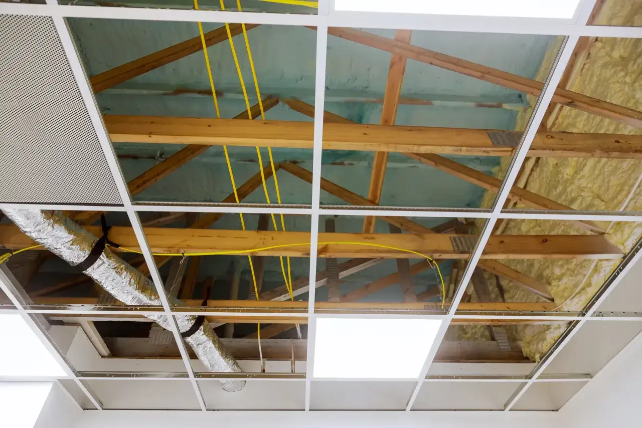 Estructura de un falso techo con vigas de madera, aislamiento, conductos y cables amarillos visibles.
