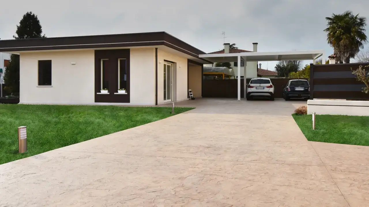 Entrada de una casa moderna con c&eacute;sped verde y camino de hormig&oacute;n cepillado. Dos coches aparcados bajo un porche.