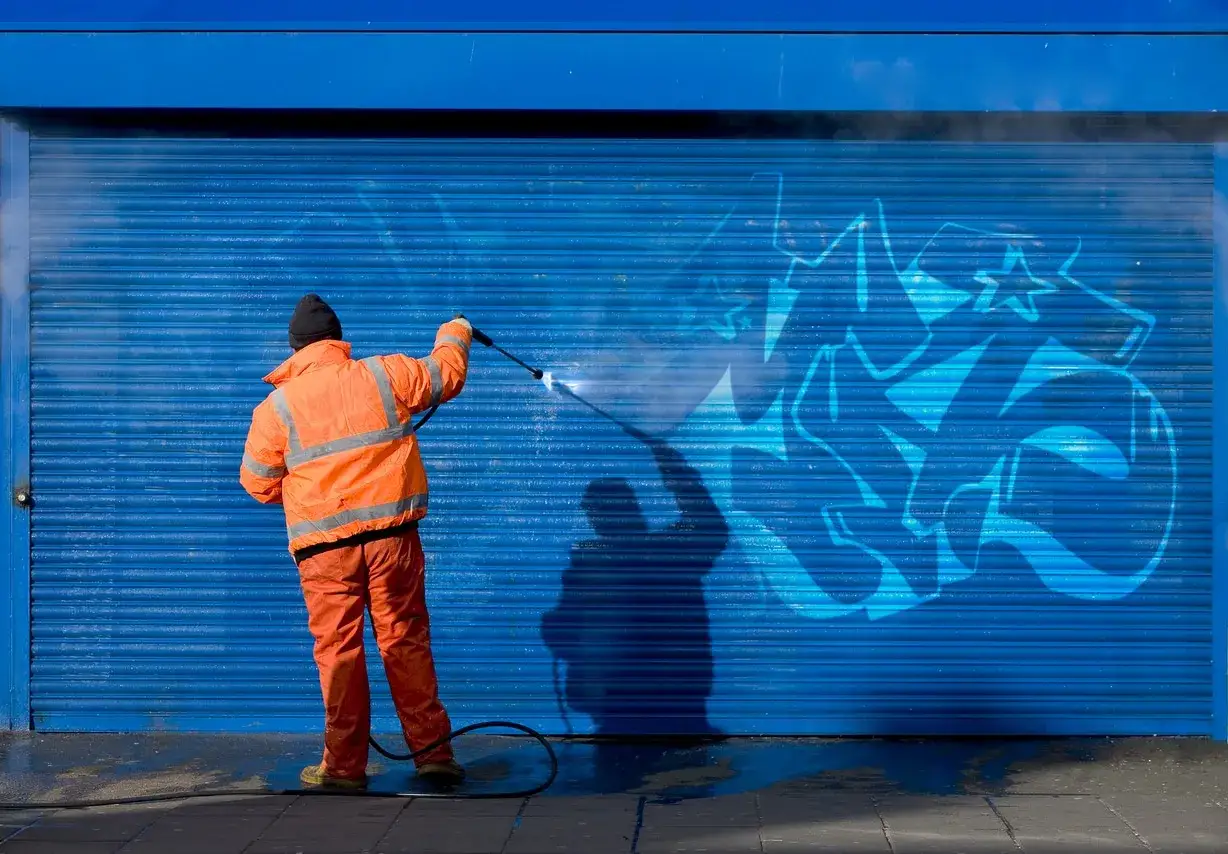 Hombre con traje naranja realiza limpieza de grafitis en una persiana azul con hidrolimpiadora.
