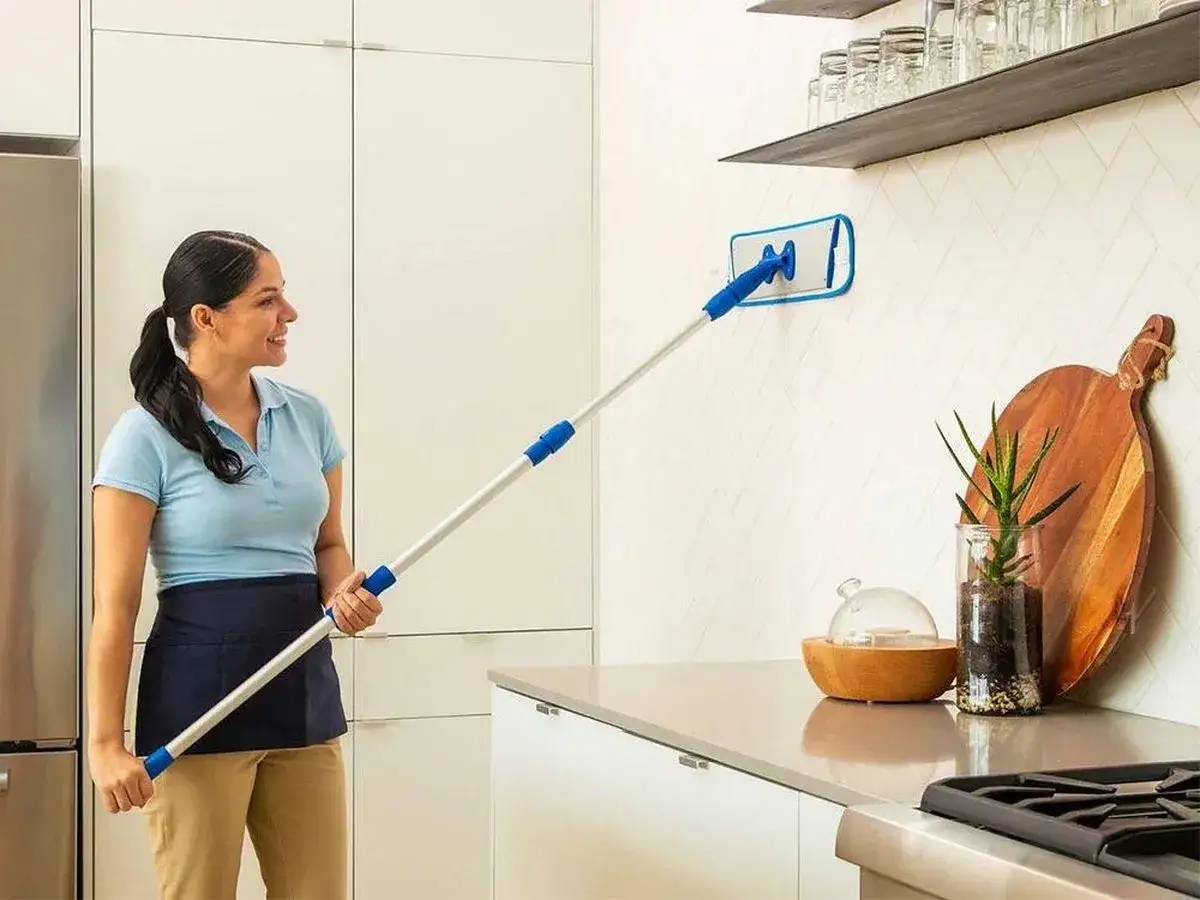 Mujer sonriente limpia azulejos con un limpiador de mango largo. El alcohol de limpieza para azulejos deja la pared impecable.