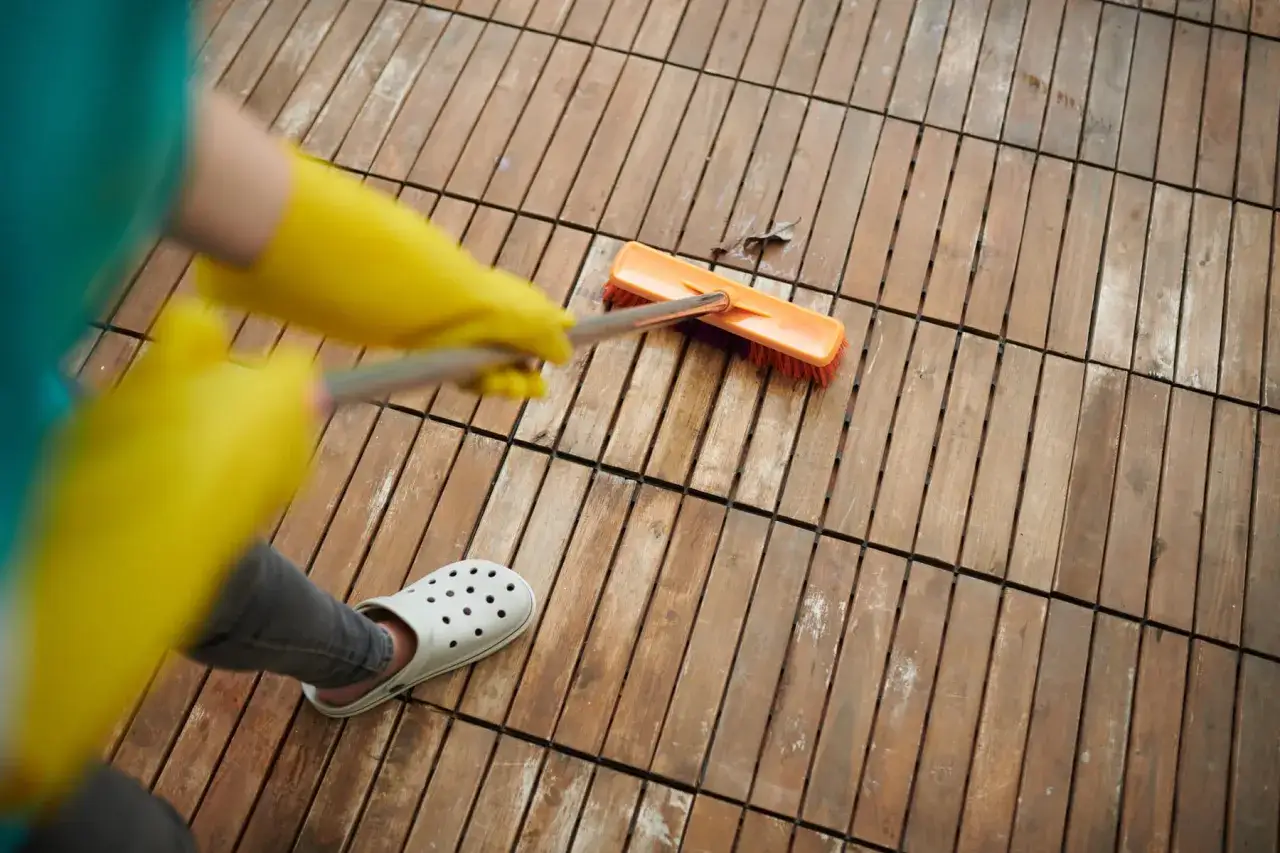 Manos enguantadas con guantes amarillos usan un cepillo para quitar manchas de vinagre en el suelo de madera.