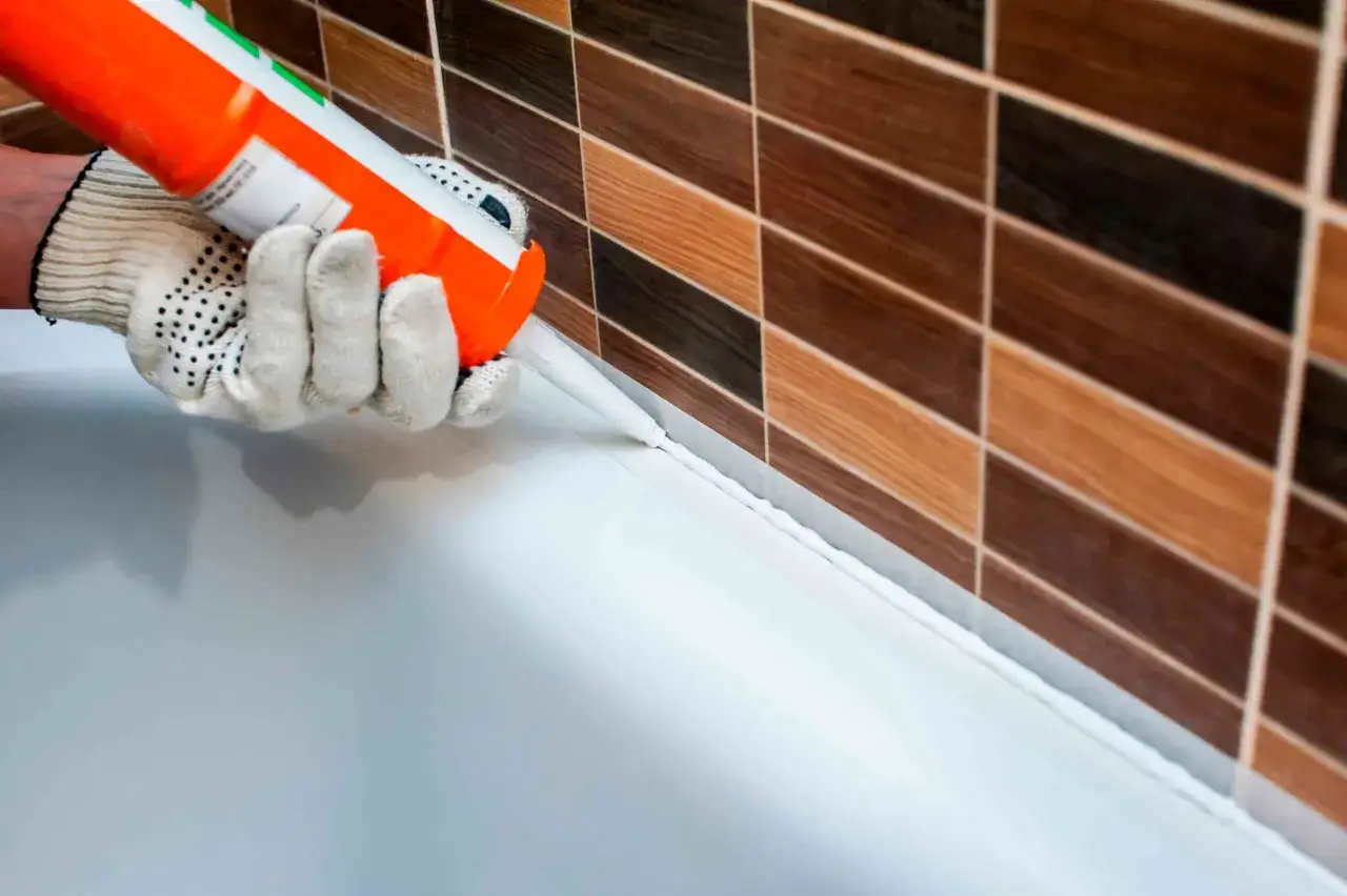 Persona con guantes aplica sellador blanco para sellar plato de ducha contra azulejos de madera.