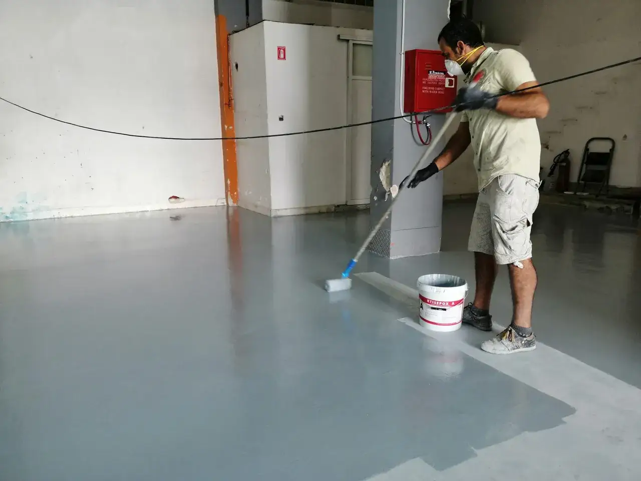 Hombre aplicando resina epoxi en suelos con rodillo. Cubo de KTISEPOX A a su lado.