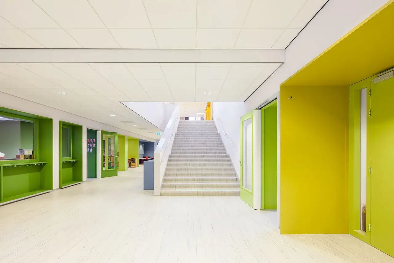 Pasillos luminosos y modernos de un colegio, con escaleras centrales, puertas verdes y amarillas, y un suelo de colegio claro.
