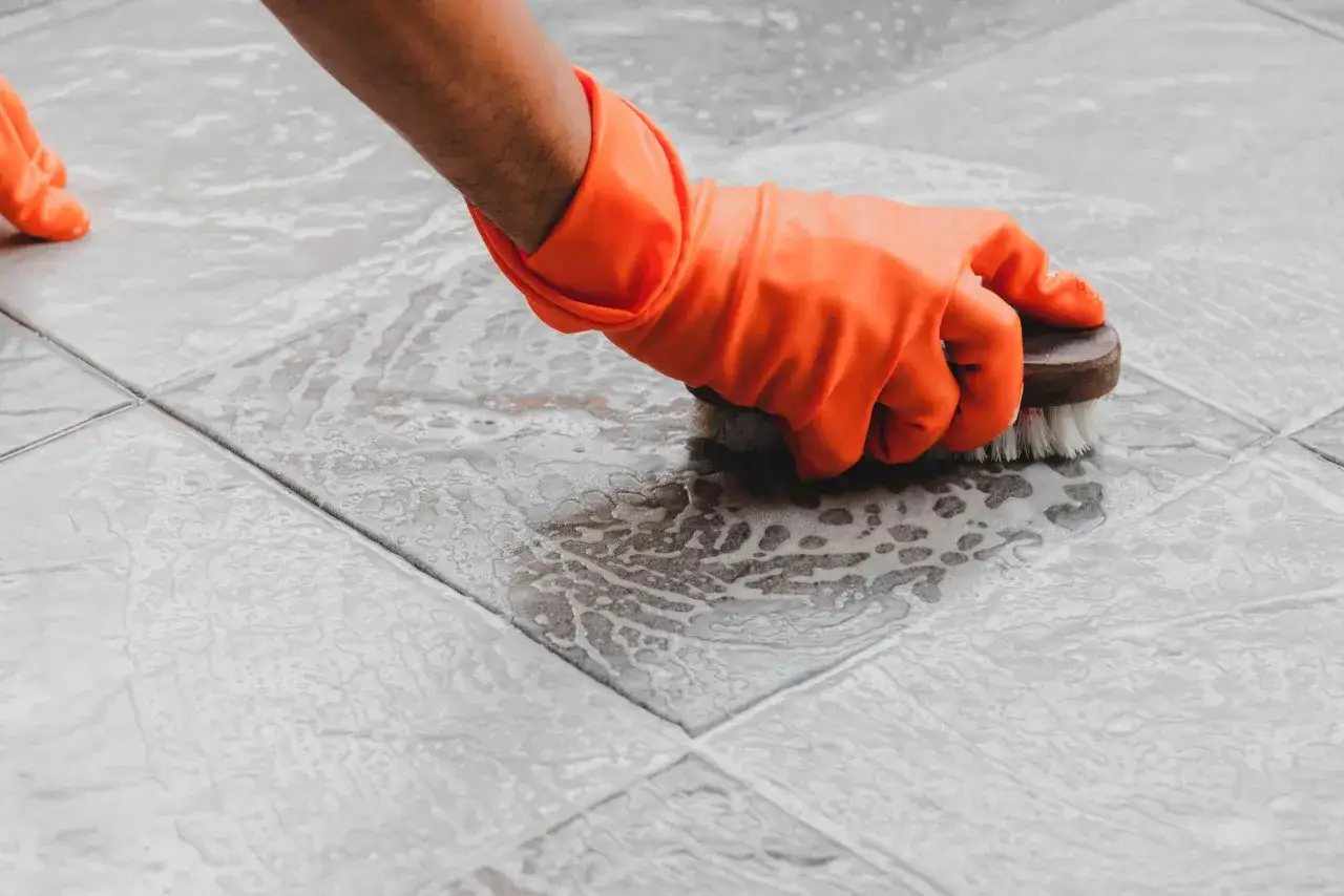 Mano con guante naranja limpiando azulejos del ba&ntilde;o con espuma y cepillo.