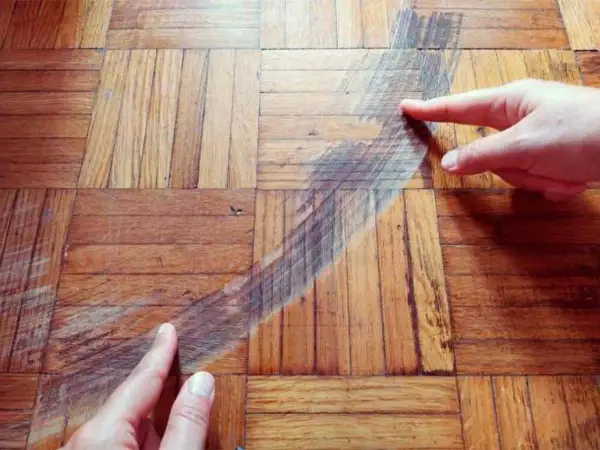 Manos señalan una mancha oscura en un suelo de parquet de madera. Se muestra cómo disimular manchas en parquet.