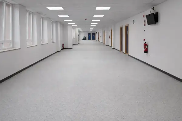 Pasillo largo y luminoso de un colegio, con ventanas a la izquierda y puertas a la derecha. El suelo del colegio es de un material gris moteado.