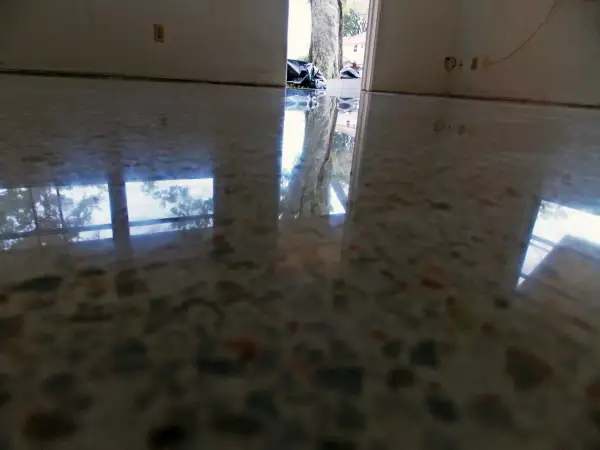 Reflejos en suelo de terrazo pulido, mostrando la entrada a una casa con bolsas de basura y un árbol. Se observa humedad en suelo de terrazo.