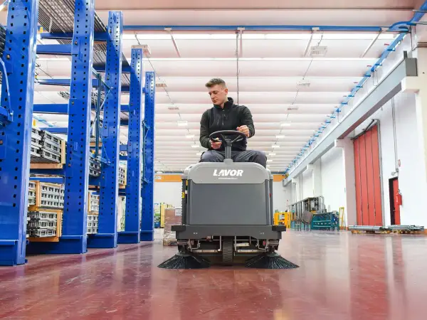 Hombre operando una maquina limpieza industrial Lavor en un almacén con estanterías azules.