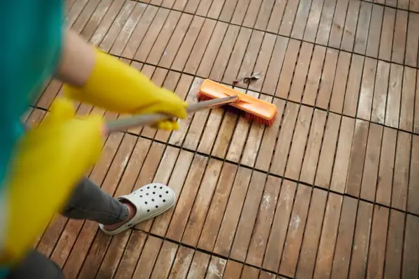 Persona con guantes amarillos y chanclas blancas usa cepillo para quitar manchas de vinagre en el suelo de madera.