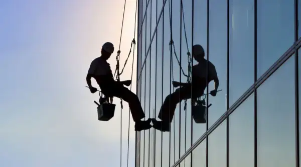 Siluetas de operarios realizando limpieza de cristales en edificios y locales, suspendidos por cuerdas.
