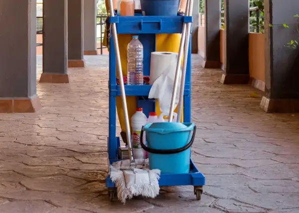 Carro de limpieza con cubeta azul, botellas y escoba sobre un suelo poroso de piedra.