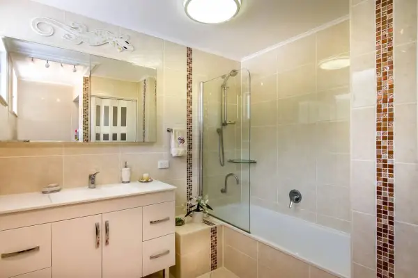 Baño con azulejos beige y detalles de mosaico. Un lavabo blanco, espejo y ducha con mampara de cristal. Ideal para aprender como poner azulejos en un baño.