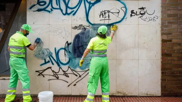 Dos trabajadores con uniformes verdes brillantes realizan la limpieza de grafitis en una pared de mármol.