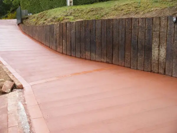 Camino de hormigón cepillado con borde de ladrillo, flanqueado por un muro de traviesas de madera.