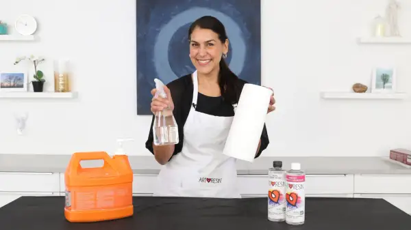 Mujer sonriente muestra cómo limpiar la resina epoxi de los utensilios con spray y papel.