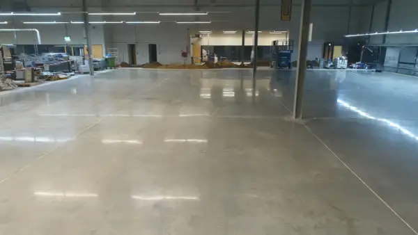 Amplio espacio industrial con suelo de **cemento pulido** impecable, reflejando las luces del techo. Se aprecian montones de tierra y maquinaria en el fondo.