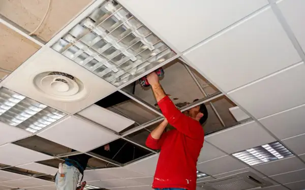 Un hombre con camisa roja instala un panel en **techos registrables**, junto a luminarias y rejillas de ventilación.