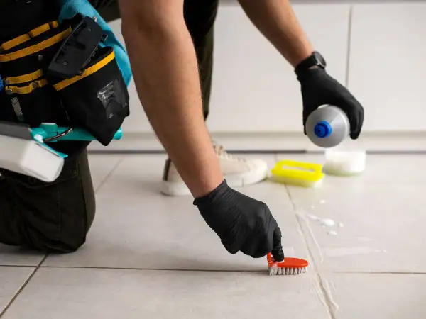Persona con guantes negros usa un cepillo para limpiar juntas de azulejos, aplicando producto de limpieza.